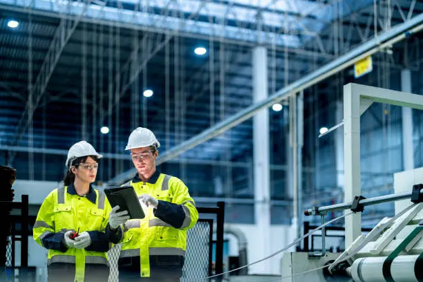 Engenheiros com capacete e colete de segurança usando tablet para monitorar processos em fábrica moderna.